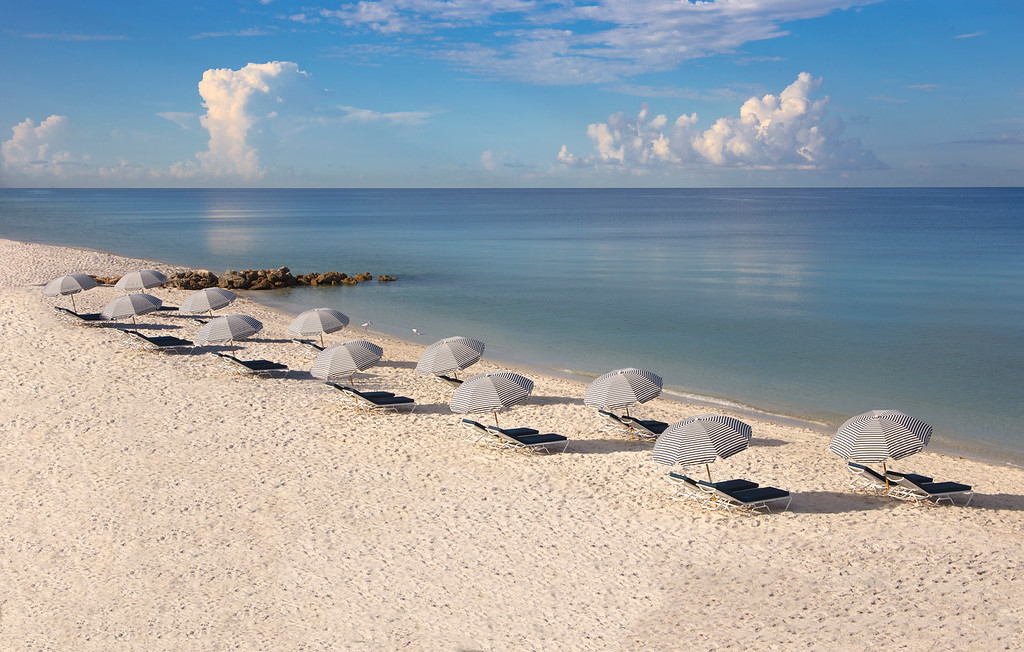 White sand beach at Edgewater Beach Hotel in Naples, with striped umbrellas, lounge chairs, calm Gulf water, and blue sky.