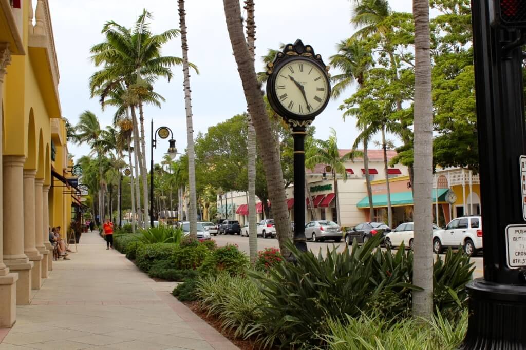 Fifth Avenue South in Naples, Florida, with palm-lined sidewalks, storefronts, parked cars, and a decorative street clock.