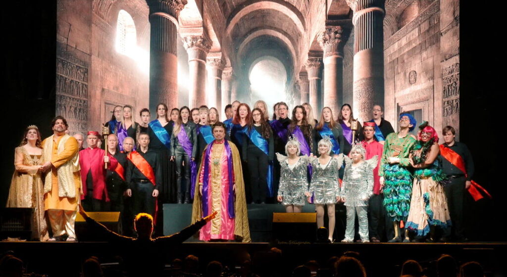 Opera cast and chorus in colourful costumes performing on stage before a grand architectural backdrop.