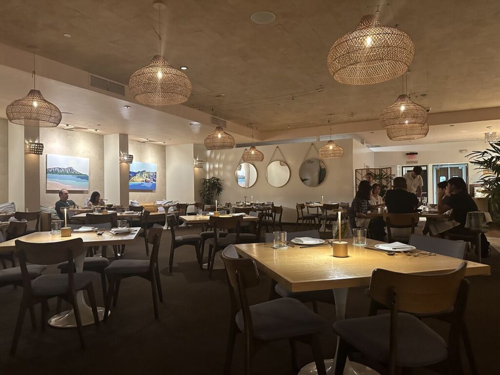 Warmly lit Arden Waikīkī dining room with woven pendant lamps, wooden tables, and island artwork.