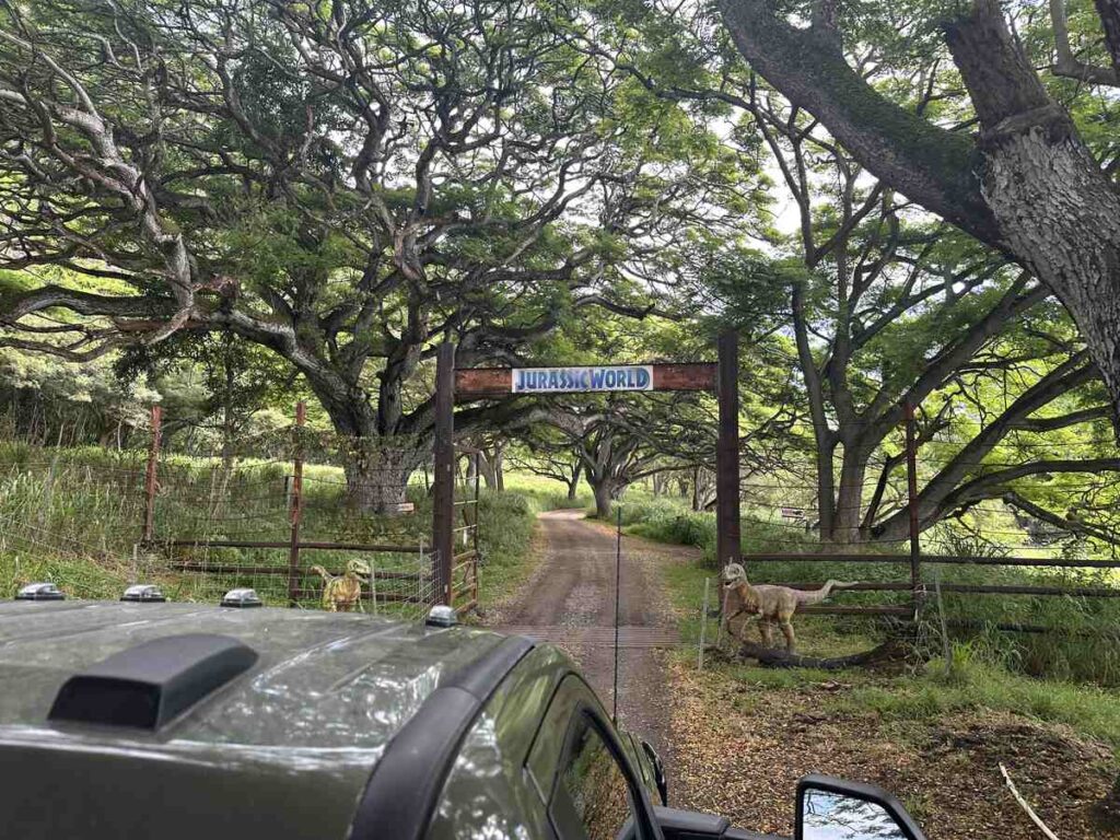 A tour vehicle approaches the Jurassic World gate beneath a canopy of trees.