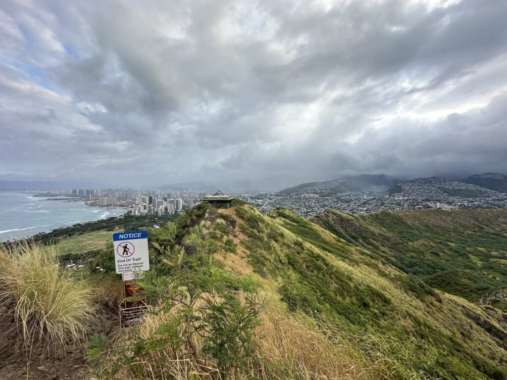 View from the Diamond Head trail over Waikīkī, Honolulu, green crater slopes, and cloudy sky.