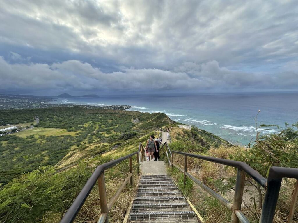 Stairs on the Diamond Head trail overlooking green crater slopes, ocean, and cloudy sky.