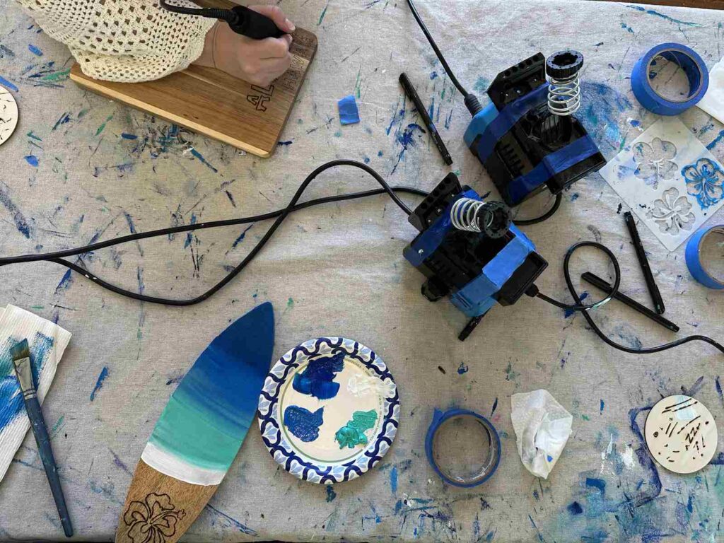 Mini surfboard art project with paint, wood-burning tools, and ocean colours on a craft table.