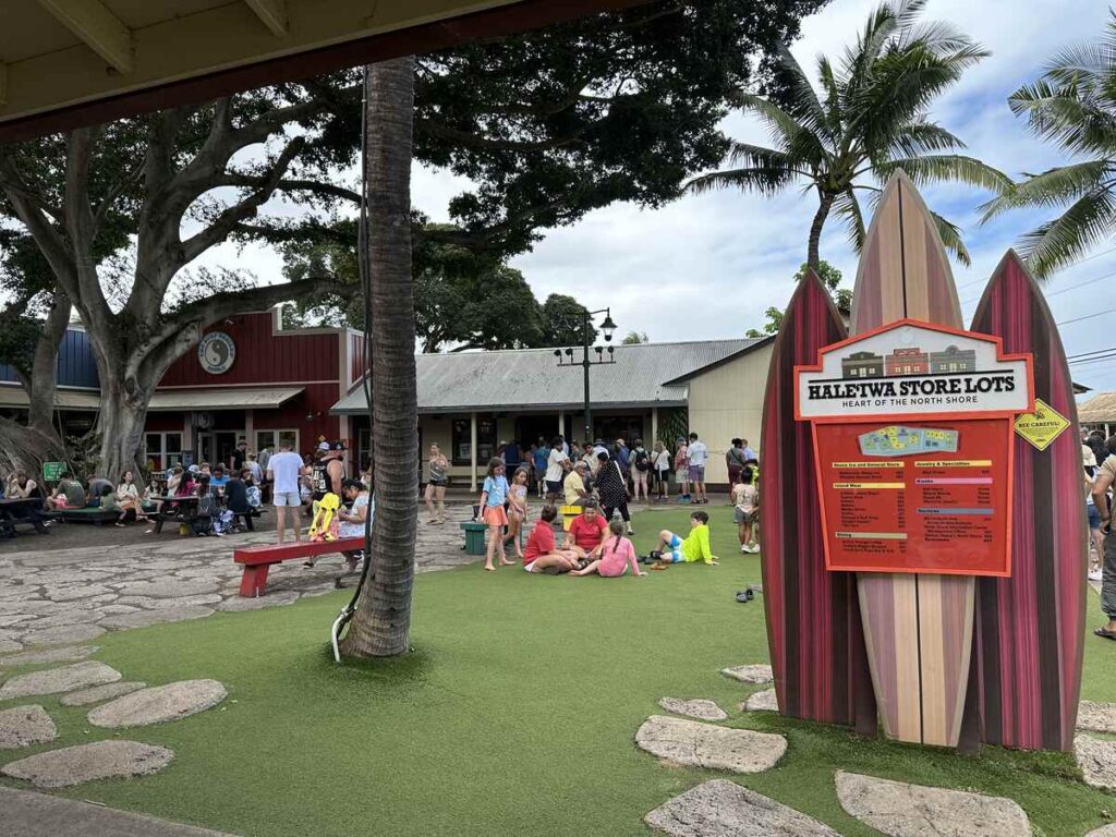 Haleʻiwa Store Lots sign shaped like surfboards with people gathered in an outdoor shopping area.