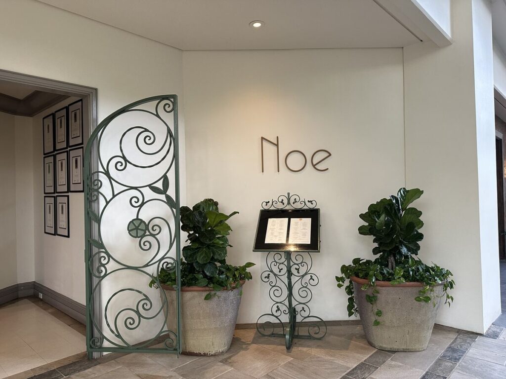 Entrance to Noe restaurant at Four Seasons Resort Oahu with potted plants, menu stand, and decorative gate.