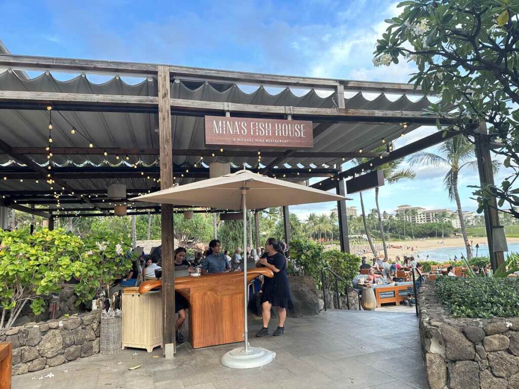 Mina’s Fish House entrance with oceanfront seating, palm trees, and beach views at Four Seasons Resort Oahu.
