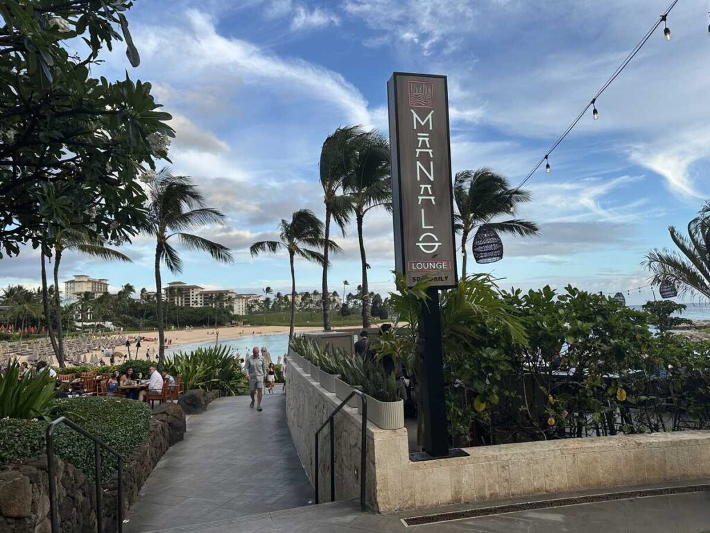 Mānalo Lounge sign beside a beach path, palm trees, string lights, and Ko Olina lagoon views.