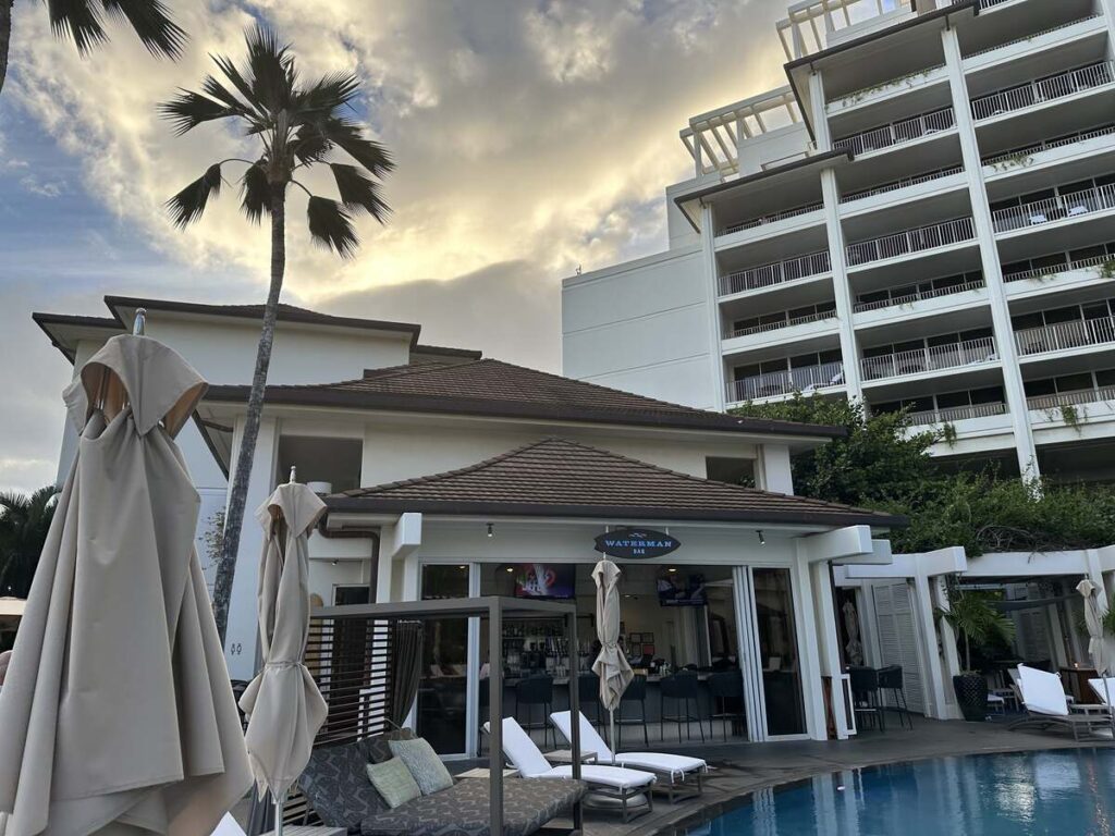 Waterman Bar beside the pool at Four Seasons Resort Oahu with lounge chairs, umbrellas, palm tree, and resort tower.