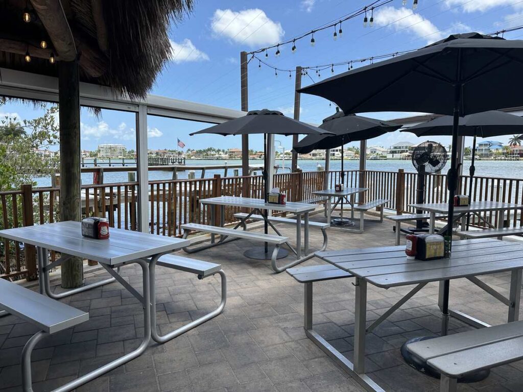 Outdoor waterfront dining patio with picnic tables, umbrellas, string lights, and views across the water in Marco Island.