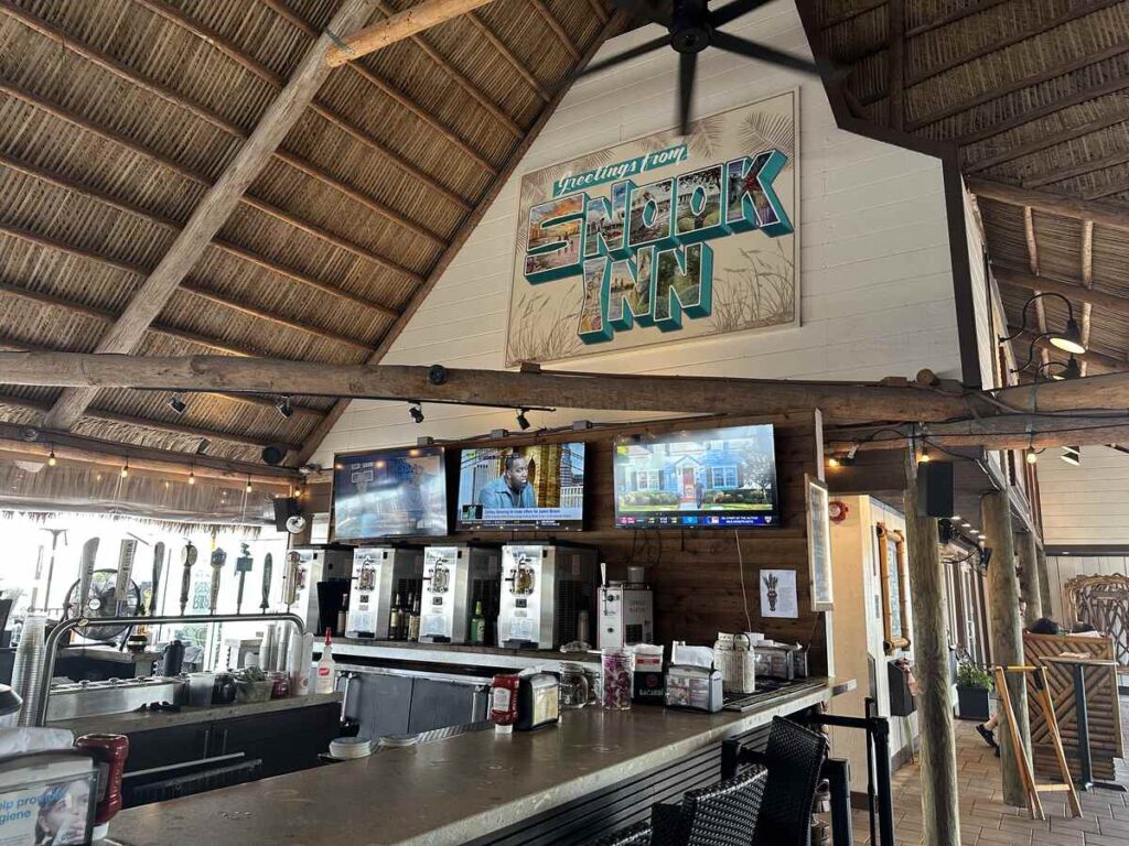 Interior bar at Snook Inn in Marco Island, with thatched roofing, wood beams, televisions, and a “Greetings from Snook Inn” sign.