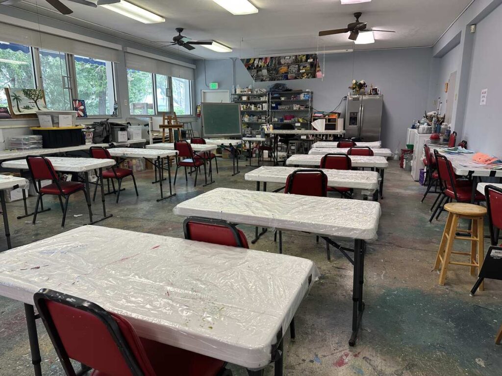 Art classroom with plastic-covered worktables, red chairs, supplies, and easels.