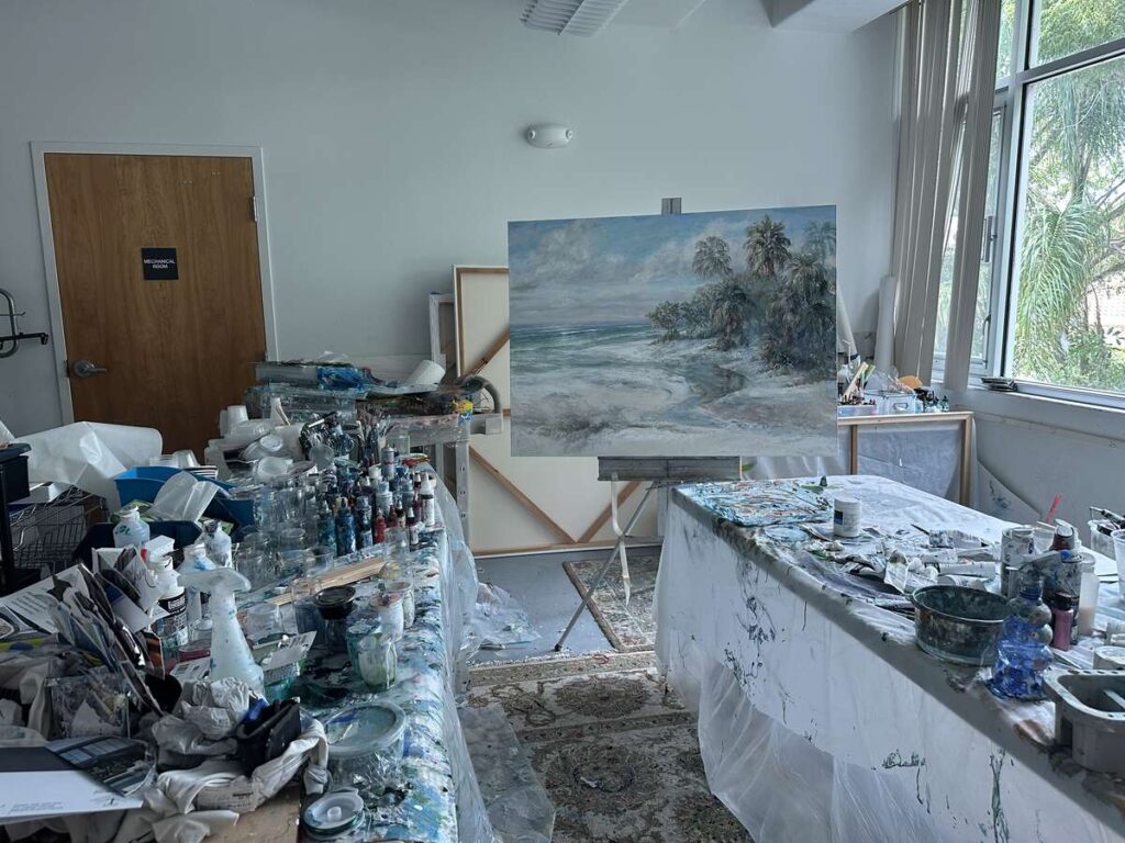 Artist Emily James’s studio at Marco Island Center for the Arts, filled with paint supplies and a coastal landscape on an easel.