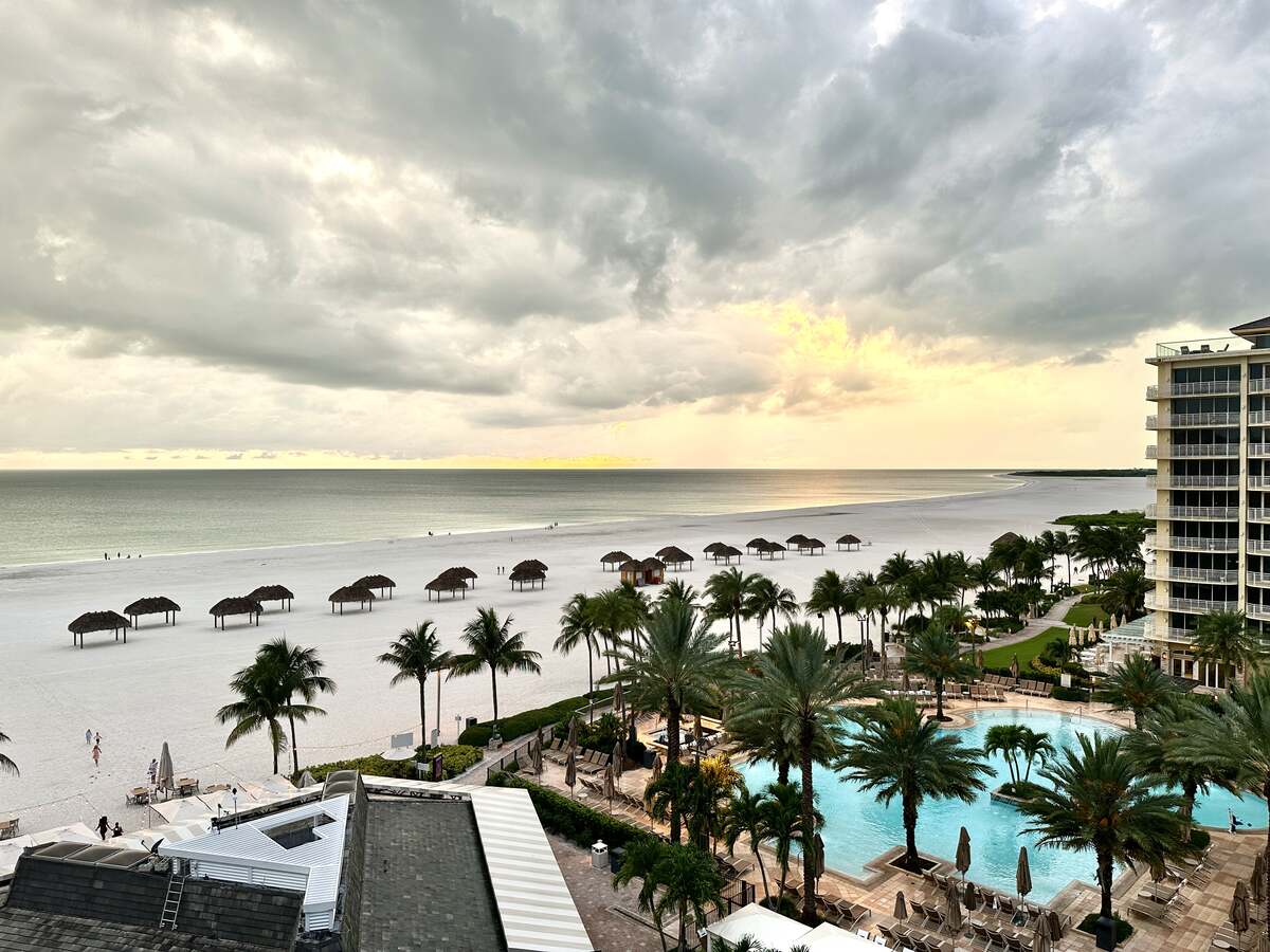 View from JW Marriott Marco Island showing a wide white-sand beach, palm trees, pool area, and dramatic sky.