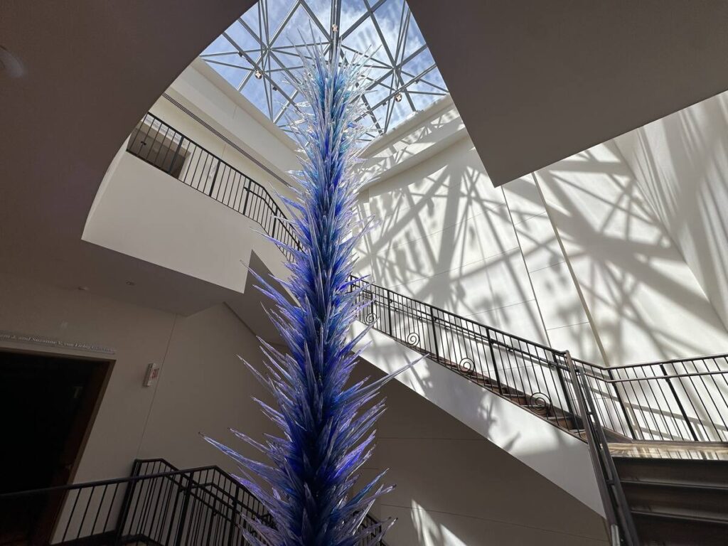 Tall blue Dale Chihuly glass sculpture rising through a sunlit stairwell at The Baker Museum.