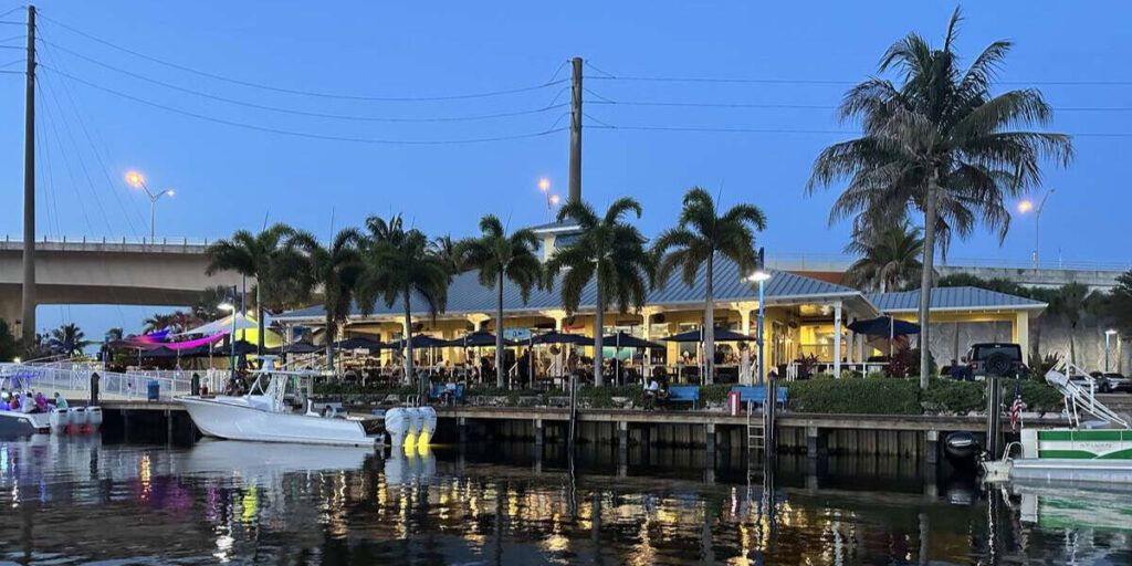 Waterfront restaurant and marina in Stuart, Florida, lit up in the evening with boats, palm trees, and reflections on the water.