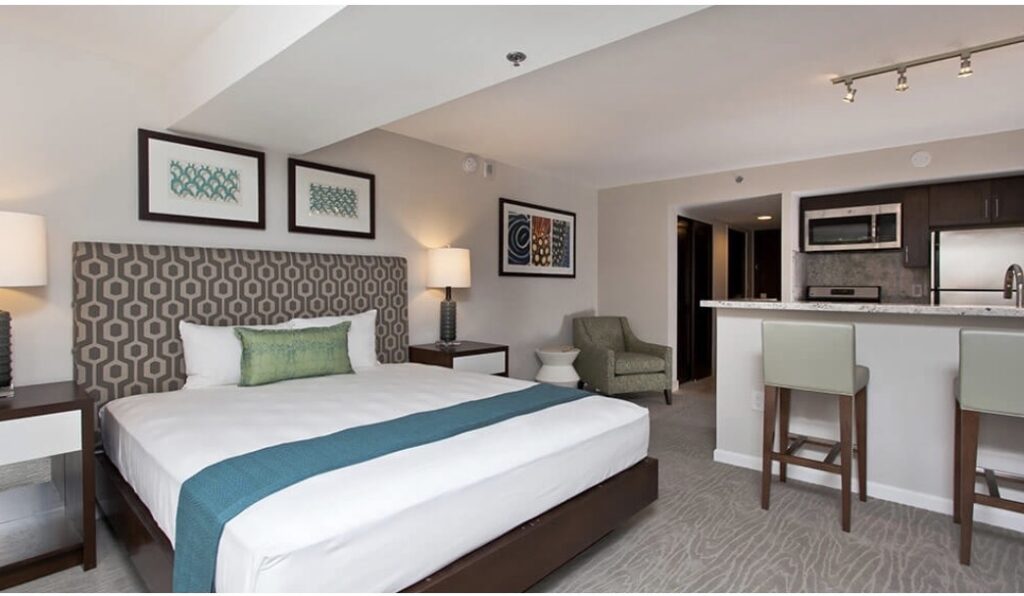 Hotel suite with a king bed, kitchenette, bar seating, and neutral island-inspired décor.