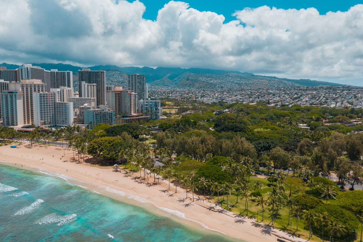 Aerial view of Waikīkī Beach, turquoise water, palm trees, hotels, and the Honolulu skyline.