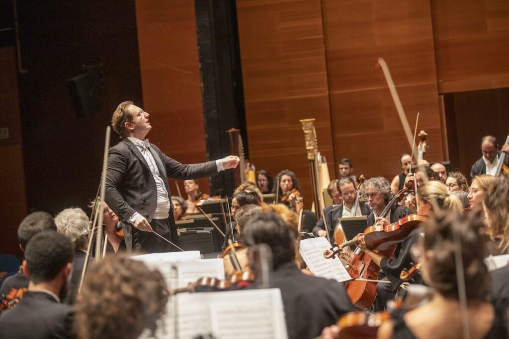 Conductor leading an orchestra during a live performance, surrounded by string musicians and music stands.