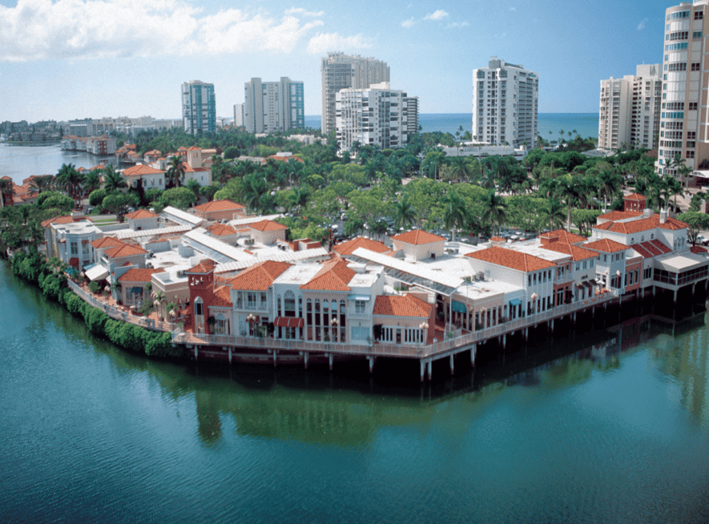 Aerial view of The Village Shops on Venetian Bay in Naples, Florida, with waterfront buildings, red-tile roofs, surrounding high-rises, and the Gulf in the distance.