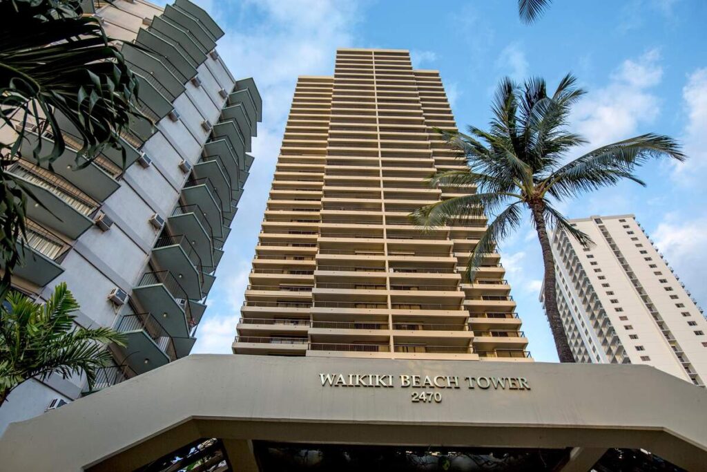 Exterior view of Aston Waikīkī Beach Tower rising above its entrance sign and palm trees in Waikīkī.