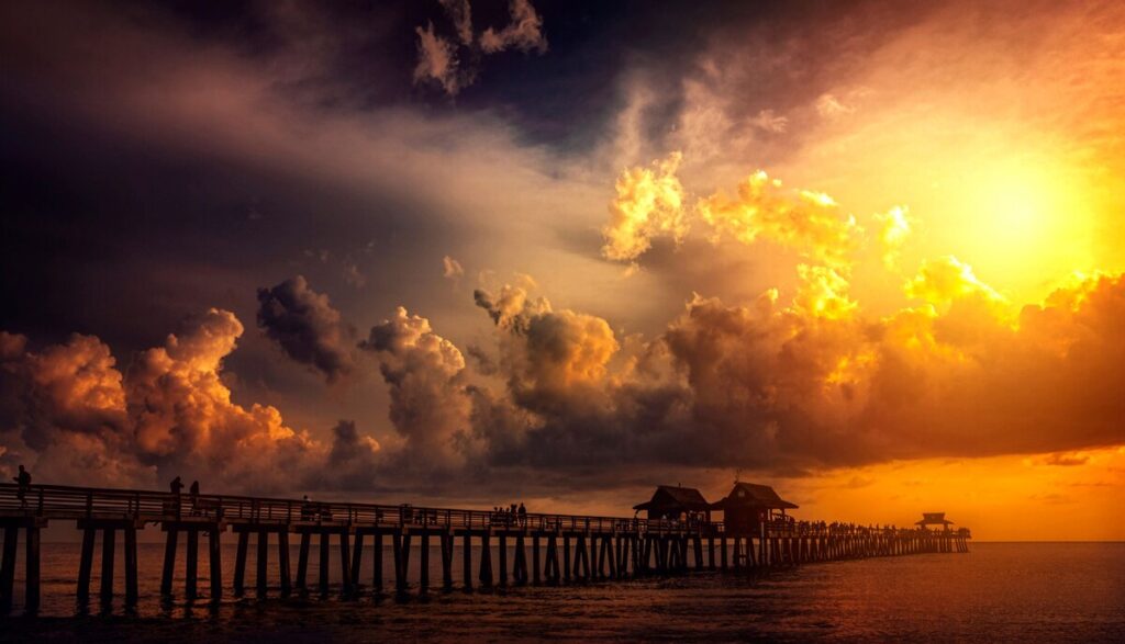 Naples Pier at sunset, with silhouetted people on the pier beneath dramatic golden clouds over the Gulf of Mexico.
