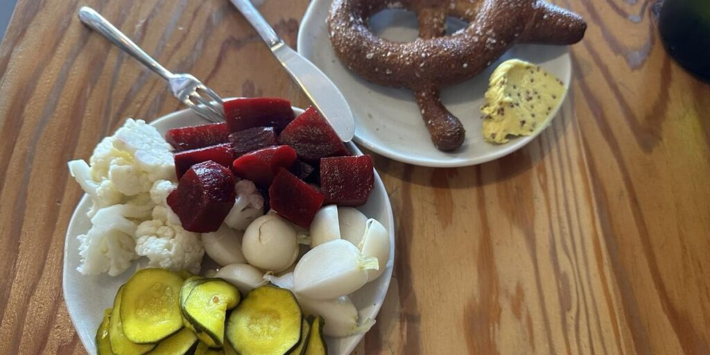 A shared plate of colourful pickled vegetables including sliced cucumbers, beets, cauliflower and onions sits beside a large soft pretzel sprinkled with coarse salt and served with mustard on a wooden table at Eva Schnitzelhaus.