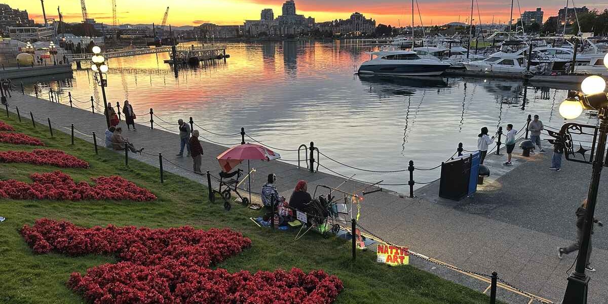 Sunset reflecting across Victoria’s Inner Harbour beside red flower beds and marina boats.